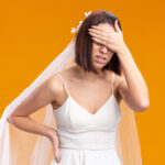 bride in beautiful wedding dress looking unwell covering eyes with hand standing over orange background