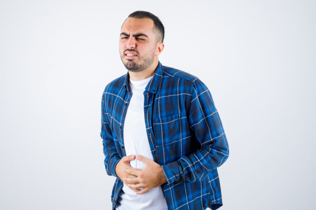young man having bellyache in checked shirt and white t-shirt and looking exhausted , front view.